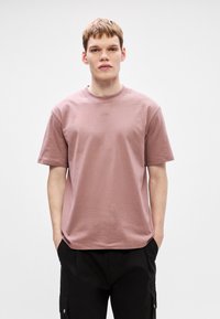 Světle růžové tričko s kulatým výstřihem vyrobené z bavlny, s krátkými rukávy a decentním logem uprostřed, nošeno s černými kalhotami.