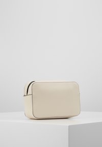 Kremowa torba z eko skórki o prostokątnym kształcie, gładkiej fakturze, z czarnym zamkiem błyskawicznym i przeszywanymi akcentami, umieszczona na białym podwyższeniu.