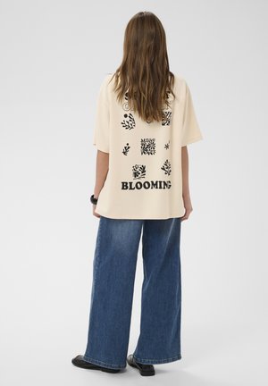 Femme aux longs cheveux bruns de dos, portant un t-shirt oversize crème avec des imprimés floraux noirs et le texte "BLOOMING", un jean large bleu et des chaussures noires.