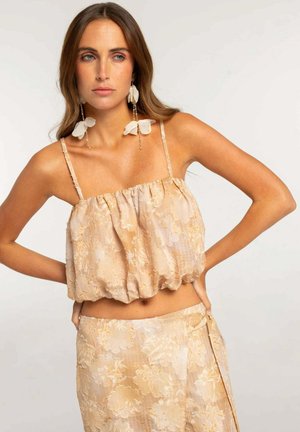 Mujer con pelo largo y castaño que lleva un conjunto beige floral de dos piezas y grandes pendientes blancos en forma de flor, de pie con las manos en las caderas.