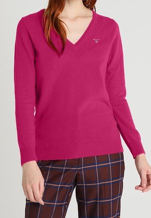 Femme portant un pull col en V rose vif et un pantalon marron à motif à carreaux bleu et orange, debout devant un fond uni.