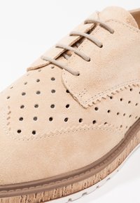 Chaussure beige en daim à lacets avec détails perforés et motifs cousus, avec une semelle texturée marron clair.