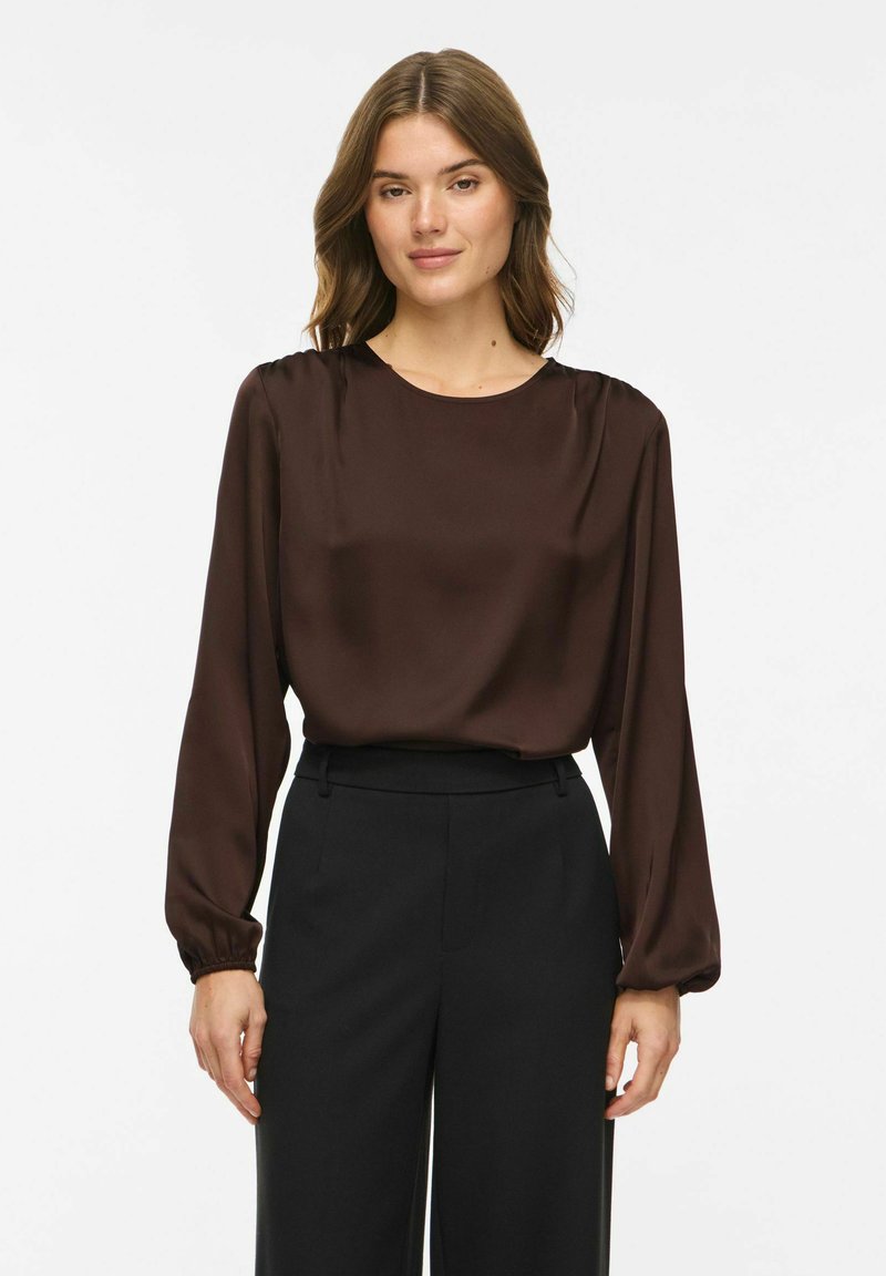 Femme portant un chemisier en satin marron foncé à manches longues, rentré dans un pantalon noir taille haute sur un fond blanc uni.