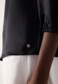 Zwarte satijnen blouse met geplooide mouwen. Voorzien van een discrete merklogo-knop aan de zoom, gecombineerd met lichte broek.