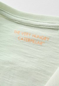 Gros plan sur un tissu vert clair avec un texte orange « THE VERY HUNGRY CATERPILLAR » cousu sous un col côtelé.
