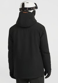 Zwarte geïsoleerde jas met een capuchon, met een gladde textuur. Subtiel "ONEILL" logo in oranje bij de hals. Gecombineerd met zwarte handschoenen en broek.