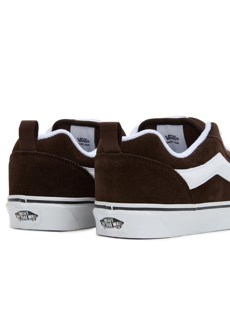 Baskets en daim marron avec des accents blancs, col rembourré, design low-top, et semelle en caoutchouc affichant le logo Vans sur le talon.