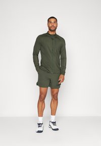 Grønn treningsjakke med glidelås og høy krage, sammen med matchende shorts. Modellen har på seg hvite og marineblå joggesko, og viser frem korte ermer og god passform.