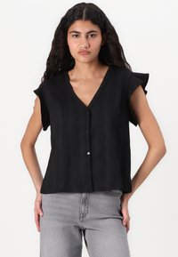 Svart kortermet topp med V-hals og knappelukking, med strukturert stoff og en avslappet passform. Kombinert med grå jeans.