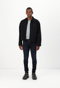 Άνδρας στέκεται με το πρόσωπο προς τα εμπρός, φορώντας ένα μαύρο φερμουάρ μπουφάν πάνω από ένα ανοιχτό γκρι πουκάμισο, σκούρο στενό τζιν και μαύρα αθλητικά παπούτσια.