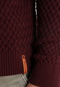 Bordeaux stickad tröja med ett texturerat rutmönster, med ribbade ärmslut och en liten orange tagg vid fållen.