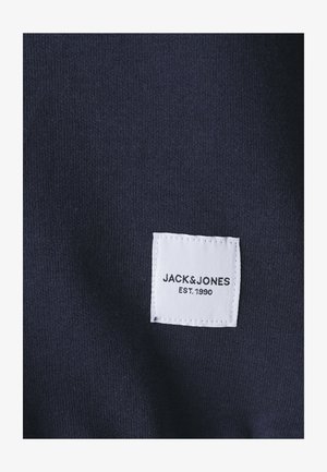 Lahja tummansininen kangas, jossa on teksturoitu neulos; valkoinen etiketti, jossa lukee "JACK & JONES EST. 1990", on ommeltu materiaaliin.