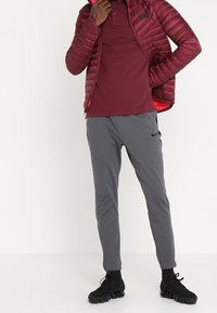 Rote gepolsterte Jacke mit Reißverschluss, kastanienbraunes Poloshirt, graue schmal zulaufende Hose und schwarze Sneaker. Glatte Textur mit minimalistischer Gestaltung.