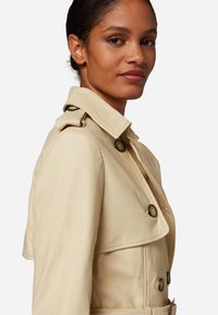 Beige läder trench coat med dubbelknäppt design, stora knappar och bred krage. Slät textur med en figurnära silhuett.