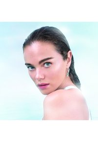 En kvinna med blött, axellångt hår, gröna ögon och en slät hy bär en silverfärgad hängörhänge och en vit linne.