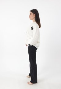 Witte gebreide trui met zwart sterpatroon, oversized fit, gecombineerd met zwarte flared jeans en naakte hakken. Zijaanzicht weergegeven.