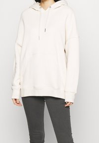 Felpa oversize color crema realizzata in tessuto morbido, con tasca frontale, cappuccio con cordini e polsini a coste, abbinata a jeans grigi scuri.