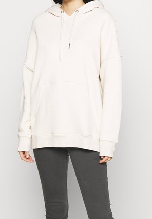 Felpa oversize color crema realizzata in tessuto morbido, con tasca frontale, cappuccio con cordini e polsini a coste, abbinata a jeans grigi scuri.