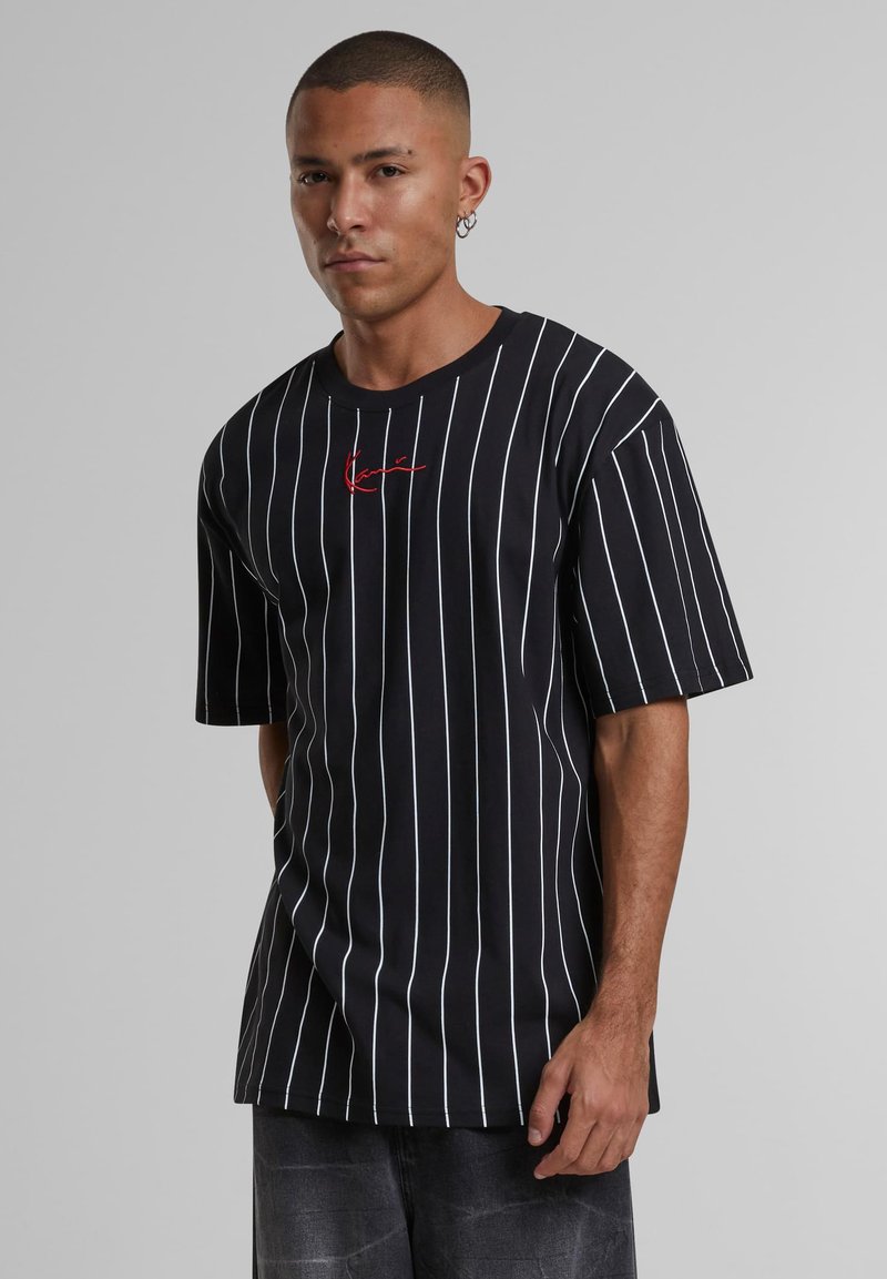 Svart T-shirt med kort ärm, vita vertikala ränder och en röd broderad logotyp på bröstet. Rund halsringning, avslappnad passform.