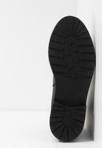 Semelle de botte noire montrant un motif de bande de roulement profond pour l'adhérence, reposant verticalement contre une surface blanche avec un coin.