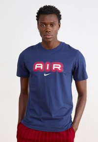 Μπλουζάκι από βαμβάκι σε ναυτικό μπλε χρώμα με μεγάλο κόκκινο και λευκό κείμενο "AIR" και λογότυπο Nike. Σχεδίαση με κοντά μανίκια και στρογγυλή λαιμόκοψη.