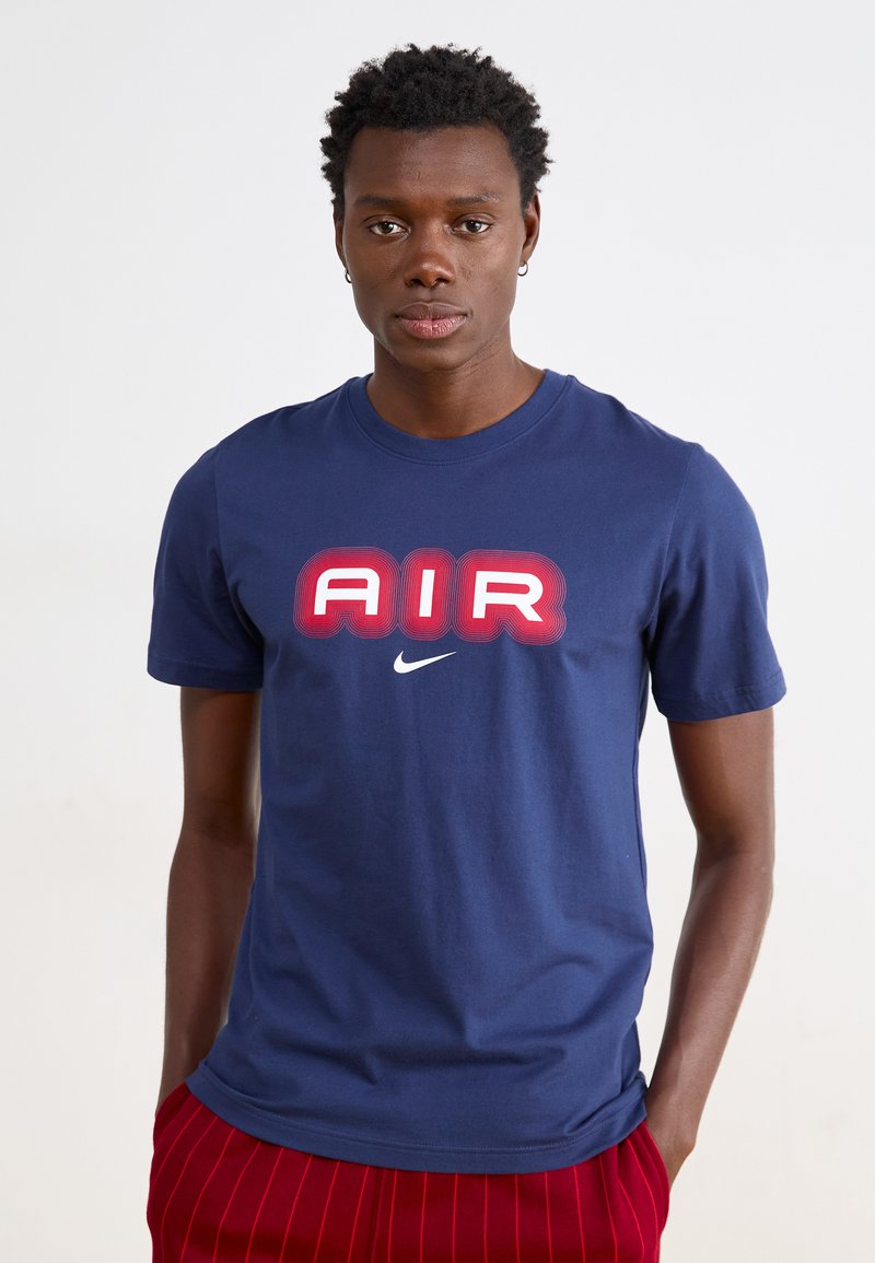Μπλουζάκι από βαμβάκι σε ναυτικό μπλε χρώμα με μεγάλο κόκκινο και λευκό κείμενο "AIR" και λογότυπο Nike. Σχεδίαση με κοντά μανίκια και στρογγυλή λαιμόκοψη.