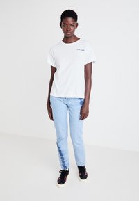 T-shirt blanc à manches courtes avec coupe décontractée, jeans bleu clair avec des patches décolorés, et baskets noires avec des accents roses.