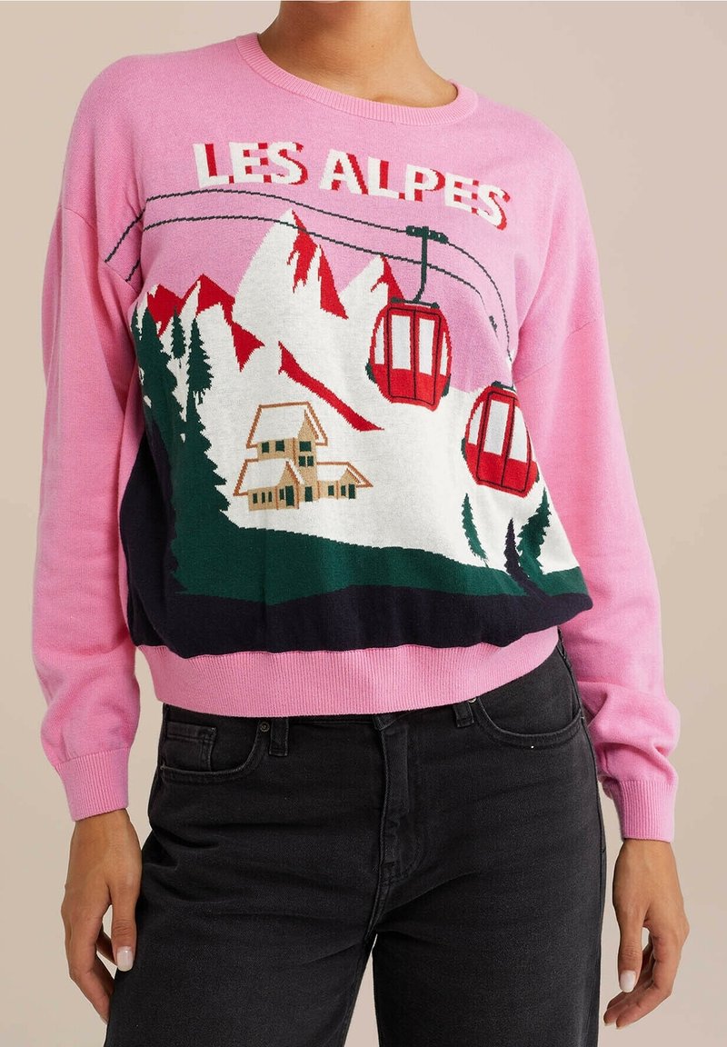 Personne portant un pull rose avec un téléphérique rouge, des montagnes, des arbres, un chalet et le texte "LES ALPES" à l’avant.