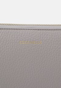 Szara skórzana saszetka o teksturowanej powierzchni, z nadrukowanym złotym logo "Coccinelle" i złotym suwakiem na górze.