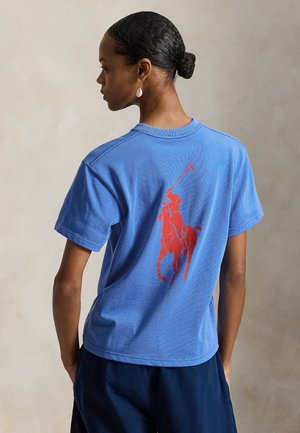 Μπλε βαμβακερό t-shirt με κόκκινο γραφικό παίκτη πόλο στην πλάτη. Κοντά μανίκια και άνετη εφαρμογή με λείο υφή.