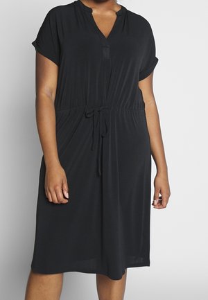 Kobieta ubrana w czarną sukienkę z krótkim rękawem, dekoltem w serek i regulowanym paskiem w talii, stojąca na gładkim jasnym tle.