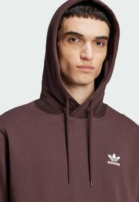Sweat à capuche bordeaux avec cordon de serrage, arborant un logo Adidas blanc sur la poitrine gauche. Fabriqué en tissu doux avec une coupe classique.