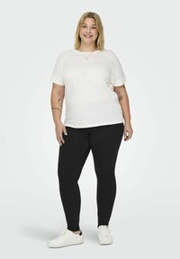 Hvid kortærmet top med teksturerede manchetter, parret med sorte skinny jeans og hvide sneakers, sat mod en lys grå baggrund.