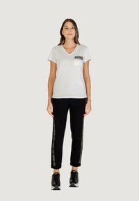 Camiseta blanca de cuello en V con un bolsillo negro y detalles impresos, combinada con pantalones deportivos negros con rayas laterales y puños enrollados. Zapatillas negras.