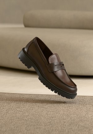 Mocassin en cuir marron foncé avec semelle épaisse en caoutchouc noir présenté au-dessus d’un tapis beige et d’un canapé en arrière-plan.