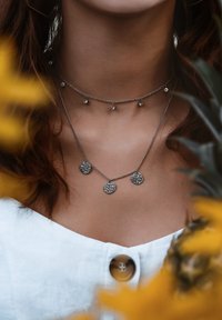 Silberne Halsketten mit strukturierten runden Anhängern und einem beaded Kettenakzent. Der Hintergrund zeigt gelbe Blumen und ein weißes Stoffoberteil.