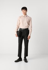 Helles rosa Langarmhemd mit subtiler Textur, schwarze Hose, Ledergürtel, schwarze Slipper und eine gefaltete schwarze Tasche in der Hand.