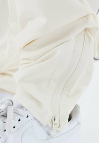 Pantalones deportivos beige con un detalle de cremallera en los tobillos, combinados con zapatillas blancas. El tejido parece suave con un ligero brillo.