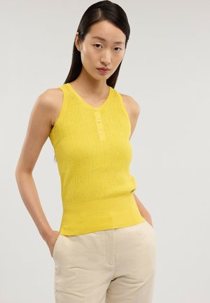 Junge Frau, die ein ärmelloses, gelbes, gestricktes Oberteil mit Knöpfen und hellfarbige Hosen trägt, vor einem schlichten Hintergrund stehend.