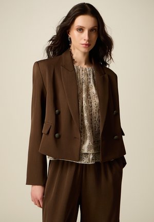 Vrouw met lang donker haar, gekleed in een bruin dubbelbreasted colbert, bijpassende broek en een beige blouse met patroon, staande voor een effen achtergrond.