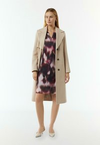 Beiger Wollmantel mit Reverskragen und doppelten Knöpfen; das Modell trägt ein plissiertes Kleid mit einem mehrfarbigen Batikmuster und hellgrauen Absätzen.