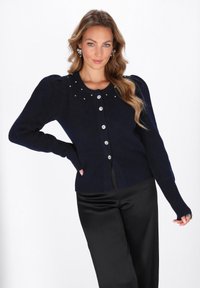Cardigan blu navy con maniche a sbuffo, decorato con borchie decorative sul colletto, con chiusura a bottoni e polsini a coste. Abbinato a pantaloni in satin neri.