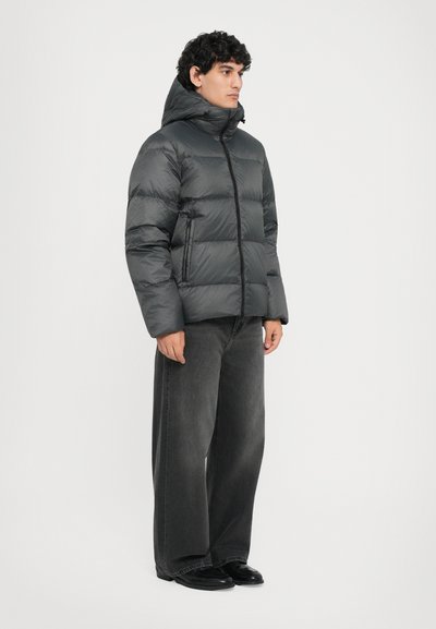 Daunenjacke in Dunkelgrau mit hohem Kragen, Reißverschluss auf der Vorderseite und horizontalen Nähten. Kombiniert mit grauen Jeans im weitem Schnitt und schwarzen Schuhen.