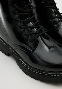 Botas de combate negras con brillo, suelas gruesas, costuras blancas detalladas y cordones negros bien ajustados sobre un fondo claro.