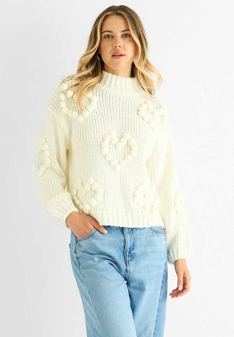 Woman wearing a white chunky knit sweater with pom-pom heart patterns, paired with light blue jeans, posing with one hand in pocket.