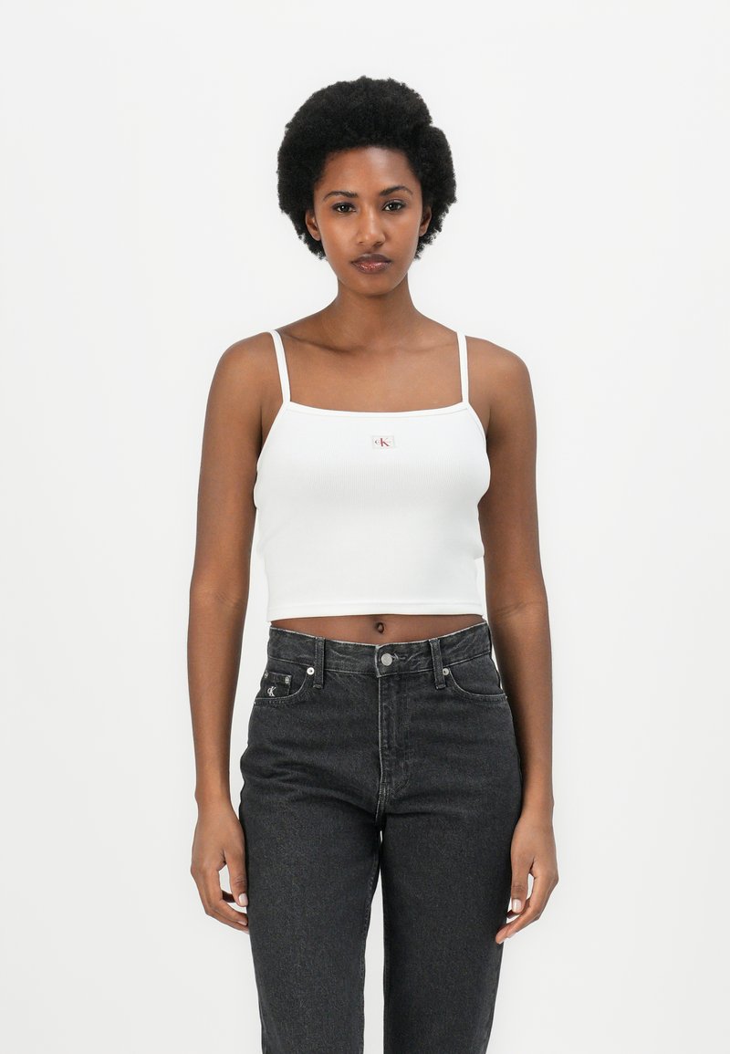 Top corto bianco a costine con spalline sottili e una piccola etichetta rossa, abbinato a jeans in denim neri a vita alta.