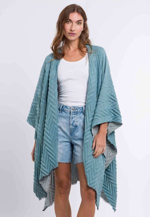 Cardigan kimono à motifs en chevrons bleu clair avec une ouverture sur le devant, en tissu texturé et aux manches larges. Associé à un short et un haut blanc.