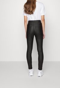 Kvinna med långt hår klädd i svarta byxor med hög midja, vit t-shirt och vita Nike-sneakers som står mot en enfärgad bakgrund.