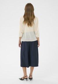 Lichtcrèmekleurige, semi-transparante blouse met geplooide driekwart mouwen, gecombineerd met marineblauwe, knie-lange, vloeiende culottes en zwarte sandalen met hak.