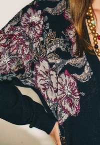 Haut noir avec broderie florale en violet et argent, à manches longues et coupe décontractée. Collier perlé ajoutant de la couleur.